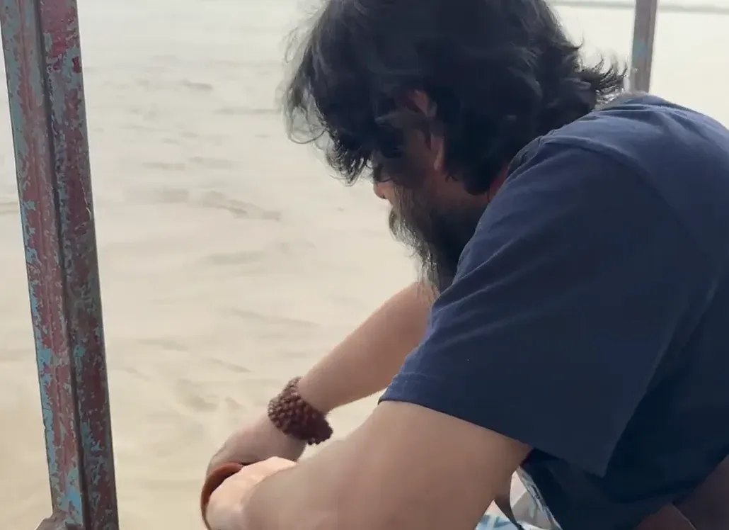 Shashwat Sanatan leaning over the rail of a boat above a silt-brown river, a rudraksha mala on his wrist.